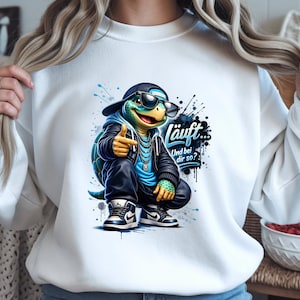 May include: White sweatshirt with a cartoon turtle wearing a black cap, sunglasses, and a leather jacket, giving a thumbs-up. The turtle is wearing a blue hoodie and sneakers. The text "läuft... Und bei dir so?" is also visible.