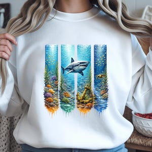 Könnte beinhalten: Weißes Sweatshirt mit einem farbenfrohen Unterwassermotiv. Die Grafik zeigt einen Hai, Korallen und einen Anker, vor dem Hintergrund blauer und grüner vertikaler Paneele, wodurch ein lebendiges und auffälliges Design entsteht.