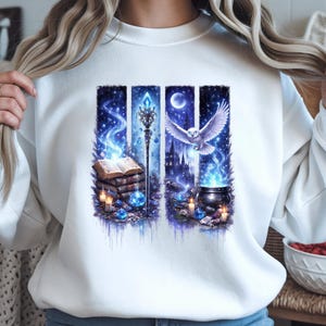 Può includere: Felpa bianca con grafica a tema fantasy. Il design include una pila di libri, un bastone, un calderone e un gufo, il tutto su uno sfondo di notte stellata con sfumature blu e viola. L'opera d'arte evoca un tema magico e mistico.