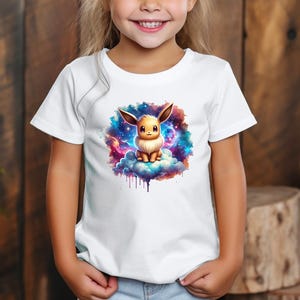 Könnte beinhalten: Weißes T-Shirt mit einer Illustration eines Cartoon-Charakters mit großen Ohren, der auf einer Wolke vor einem Galaxiehintergrund sitzt. Das Design ist farbenfroh und auffällig, ideal für Kinder.