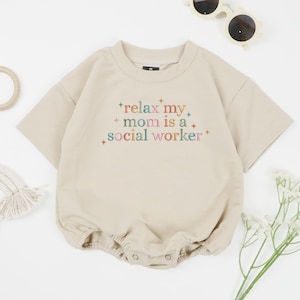 May include: A beige baby romper with short sleeves and a round neckline. The romper features the text "relax my mom is a social worker" in colorful embroidery. The image also includes sunglasses, a wooden toy, and flowers.