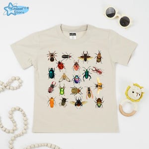 May include: Beige crew neck t-shirt featuring a colorful insect illustration. The graphic includes various beetles, bugs, and insects in a grid pattern. The shirt is styled with sunglasses and a baby toy.