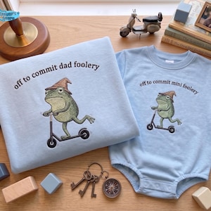 May include: Light blue sweatshirt and onesie set featuring a cartoon frog riding a scooter and wearing a hat. The sweatshirt reads "off to commit dad foolery," and the onesie says "off to commit mini foolery."