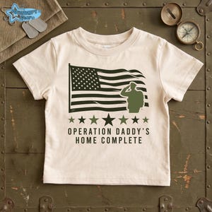 May include: Cream-colored toddler t-shirt featuring a black and white American flag graphic, a saluting soldier silhouette, and the text "OPERATION DADDY'S HOME COMPLETE." The design includes green stars.