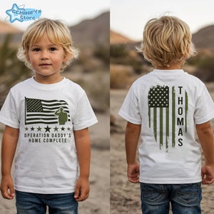 May include: White t-shirt featuring an American flag design with the text "OPERATION DADDY'S HOME COMPLETE" on the front. The back of the shirt displays a distressed American flag with the name "THOMAS" printed vertically.