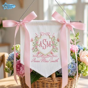 May include: A white fabric banner hanging from a metal rod with pink satin ribbons. The banner features an embroidered gold crest with the pink monogram ASM, surrounded by floral vines and butterflies, with the name Anna Mae Smith below.