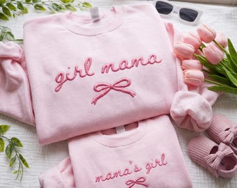 May include: Two light pink sweatshirts with embroidered text and bow details. The top sweatshirt says "girl mama" and the bottom one says "mama's girl". The image also includes pink slippers, sunglasses, and pink tulips.