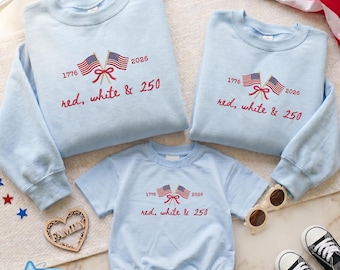 Pode incluir: Camisolas azuis claras e um body para bebé com bandeiras bordadas a vermelho, branco e azul e texto. O texto diz "1776 2026 red, white & 250". Os artigos estão dispostos com óculos de sol e sapatos.
