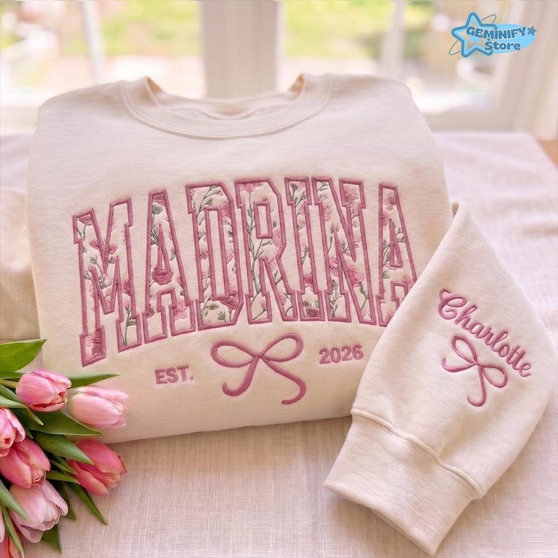May include: Cream-coloured sweatshirt with the name "MADRINA" embroidered in pink floral letters. The sleeve features the name "Charlotte" in pink script. A pink bow and the year "2026" are also embroidered on the sweatshirt.