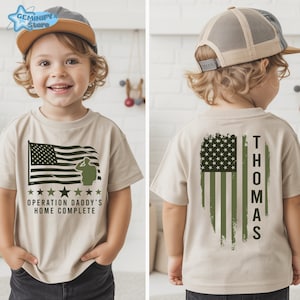 May include: Tan short-sleeved t-shirt featuring an American flag design with a saluting soldier silhouette. The text "OPERATION DADDY'S HOME COMPLETE" is below the flag. The back of the shirt displays a distressed American flag and the name "THOMAS".
