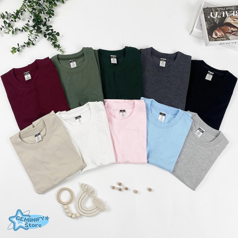May include: A selection of folded t-shirts in a range of colours, including burgundy, olive green, navy, charcoal, and light blue. Other colours are beige, white, and pink. The shirts are arranged in a fan shape on a white surface.