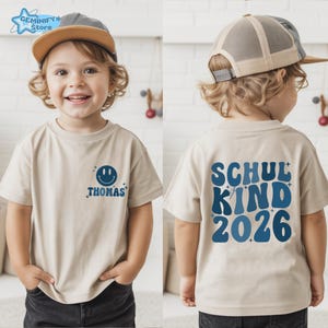 May include: Beige short-sleeved t-shirt with a blue smiley face and the name "Thomas" printed on the front. The back of the shirt features the text "SCHUL KIND 2026" in blue. The shirt is worn by a child.