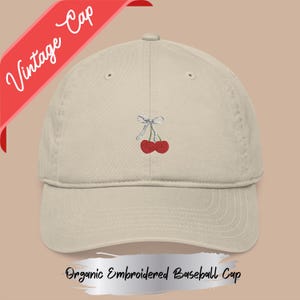 May include: A beige organic embroidered baseball cap with a red cherry design and a silver bow. The cap has the words "Vintage Cap" on a red banner. The text "Organic Embroidered Baseball Cap" is also visible.