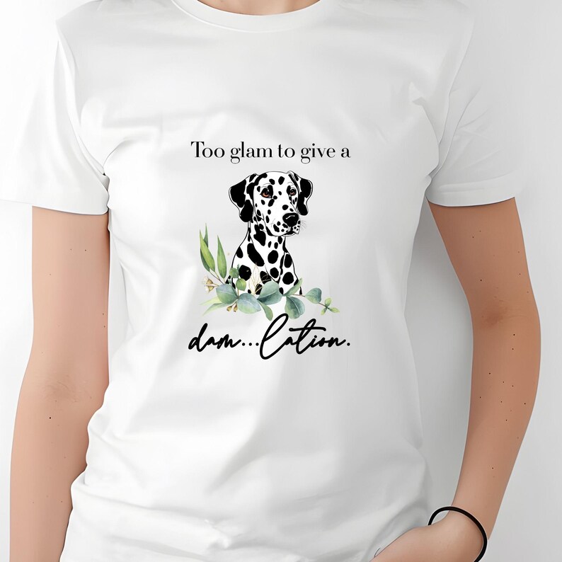 May include: White t-shirt featuring a Dalmatian dog illustration with black spots and greenery. The shirt has the text "Too glam to give a damn...lation" in black script. The shirt is a casual wear item.