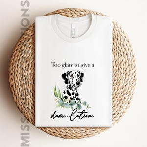 May include: White t-shirt with a graphic of a Dalmatian dog and the text "Too glam to give a damn...cation." The dog has black spots and is surrounded by green leaves. The shirt is displayed on a woven basket.
