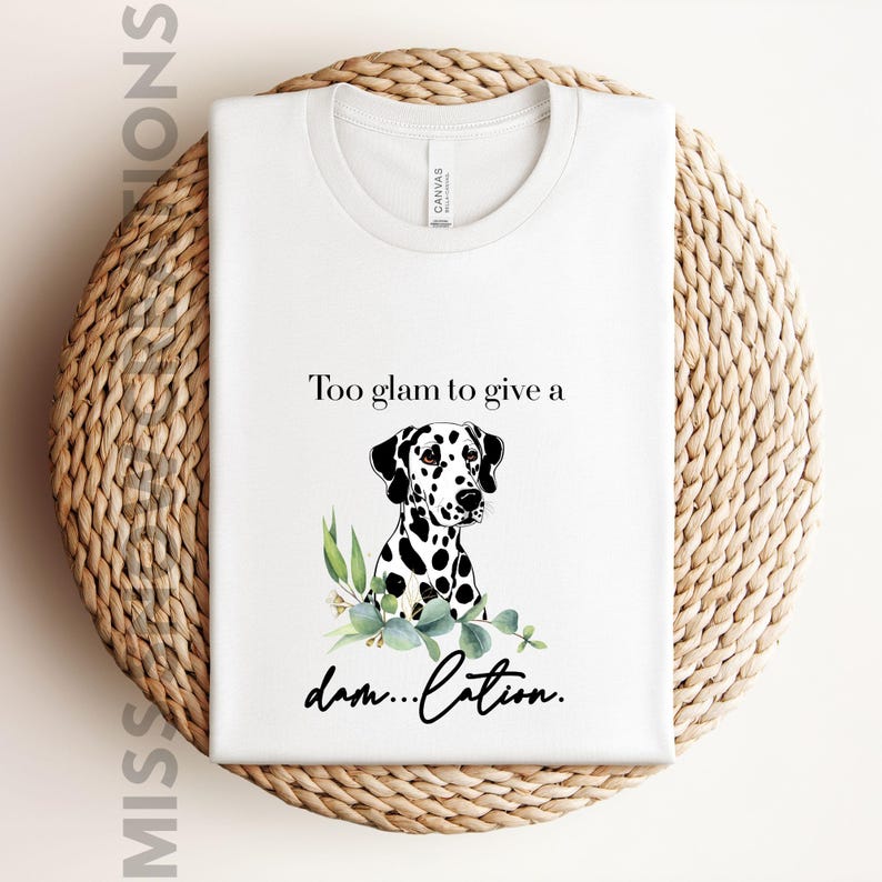 May include: White t-shirt with a graphic of a Dalmatian dog and the text "Too glam to give a damn...cation." The dog has black spots and is surrounded by green leaves. The shirt is displayed on a woven basket.