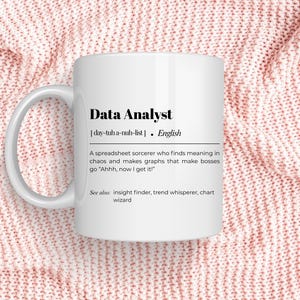 Peut inclure: Mug en céramique blanche avec le texte "Data Analyst" et une définition. La définition décrit un analyste de données comme un sorcier de tableur. Le mug est sur une surface tricotée rose.