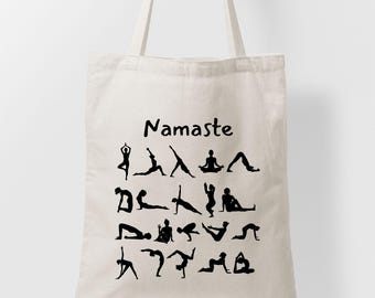 May include: Cream-colored tote bag featuring black silhouettes of yoga poses. The word "Namaste" is printed in black script above the yoga poses. The bag has two cream-colored handles.