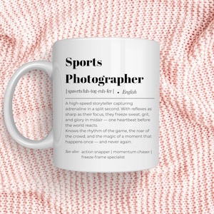 May include: White ceramic mug with the text "Sports Photographer" and a definition. The mug is on a pink knitted surface. The definition describes the role of a sports photographer.