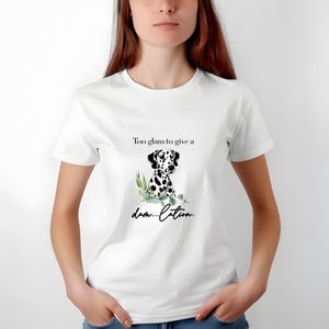 May include: White t-shirt featuring a Dalmatian dog illustration with black spots and green leaves. The text on the shirt reads "Too glam to give a damn...lation."