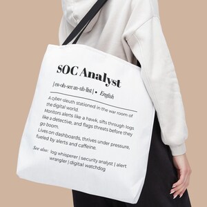 Puede incluir: Bolso tote blanco con correas negras que muestra el texto "SOC Analyst" en una fuente negra. El bolso incluye una definición del término, que describe el rol en el mundo digital. Un accesorio elegante y práctico.