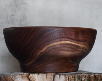 May include: A dark brown wooden bowl with a smooth, polished finish and visible natural grain patterns. The bowl features a wide, rounded upper rim that tapers down to a narrower, solid base, showcasing the rich, deep tones of the wood.