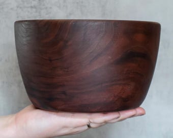 May include: A deep, rounded wooden bowl featuring a dark brown finish with visible natural wood grain patterns. The bowl has a smooth, curved exterior surface and a wide, open top, showcasing the rich, varied tones of the dark timber material.