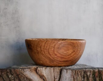 May include: A round wooden bowl with a visible natural grain pattern, featuring a warm brown tone and a smooth, polished finish. The bowl has a curved shape and a consistent rim, showcasing the organic texture of the wood material.