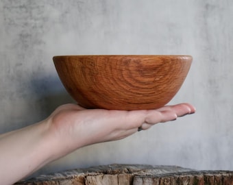 May include: A round, medium-brown wooden bowl with a visible, swirling grain pattern. The bowl has a smooth, curved exterior and a simple, open top design, showcasing the natural texture and warm, reddish-brown tones of the wood material.