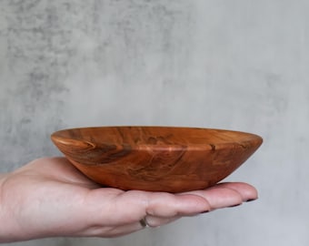 May include: A small, shallow wooden bowl featuring a rich, warm brown color with prominent, swirling natural grain patterns throughout the surface. The bowl has a smooth, rounded rim and a curved exterior profile, showcasing the organic texture of the wood.
