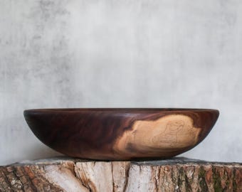 May include: A round wooden bowl featuring a deep brown, polished finish with a prominent, lighter-colored natural wood grain patch on the side. The bowl has a smooth, curved rim and a wide, shallow basin, showcasing the organic texture of the wood.