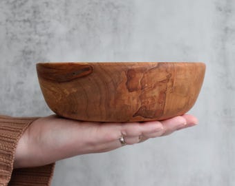 May include: A round wooden bowl with a smooth, polished finish and visible natural grain patterns in shades of light and dark brown. The bowl features organic, swirling wood textures and a curved rim, showcasing the raw beauty of the timber.