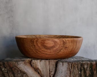 May include: A round wooden bowl with a visible natural grain pattern. The bowl features a warm, medium-brown tone and a smooth, curved exterior surface. The wood displays distinct circular growth rings and a textured finish along the outer rim.