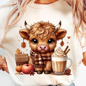 Può includere: T-shirt color crema con un'illustrazione a cartoni animati di una mucca delle Highlands. La mucca indossa una sciarpa a quadri ed è circondata da elementi a tema autunnale, tra cui una zucca, una mela e una tazza di caffè con panna montata.