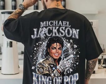 Könnte beinhalten: Schwarzes T-Shirt mit einem Porträt von Michael Jackson, dem Text "MICHAEL JACKSON" und "KING OF POP" in einer glitzernden Schriftart. Das Bild zeigt Jackson in einer Jacke und einem Handschuh mit Juwelen, lächelnd.