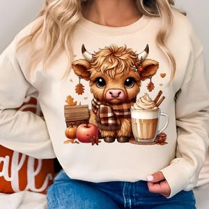 May include: Beige sweatshirt featuring a cartoon Highland cow wearing a plaid scarf, with a mug of latte, pumpkins, and fall leaves. The cow has large eyes and is surrounded by autumn-themed elements.