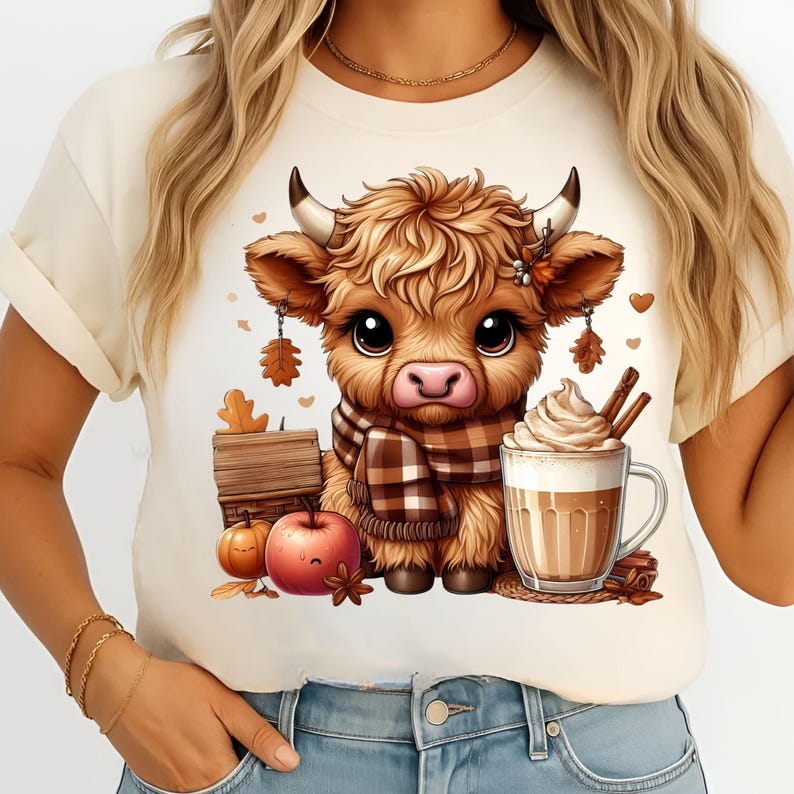 May include: Cream-colored t-shirt featuring a cartoon Highland cow with a plaid scarf, surrounded by fall-themed elements like pumpkins, apples, and a latte. The cow has large eyes and is the central focus of the design.