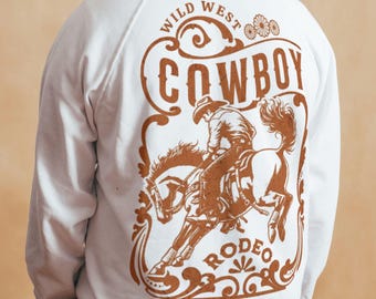 May include: White sweatshirt with a brown graphic of a cowboy riding a horse, with the words "COWBOY RODEO" and "WILD WEST" in a decorative font. The design is centered on the back of the sweatshirt.