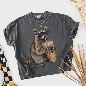 Puede incluir: Camiseta gris oscuro con un mapache con gafas de sol bebiendo de una taza de café. La taza tiene el número 73. El mapache es el gráfico principal de la camiseta.