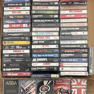 May include: A collection of numerous music cassette tapes arranged in a box, featuring various artists such as Aerosmith, The Beatles, Bon Jovi, and ABBA. Each plastic case displays the artist name and album title on the spine or front cover.