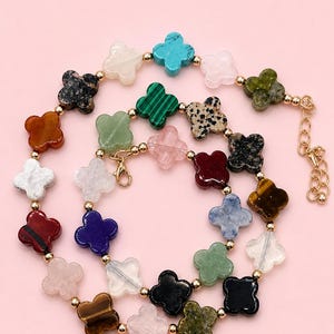 May include: A colorful necklace featuring clover-shaped beads in various colors and textures, including turquoise, green, and red. The beads are strung together with gold-colored metal accents and a clasp. The necklace is displayed against a pink background.