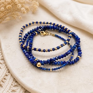 May include: A stack of three beaded bracelets. The bracelets feature deep blue lapis lazuli beads and small gold-colored beads. A gold-colored clasp secures the bracelets. The bracelets are displayed on a white surface.