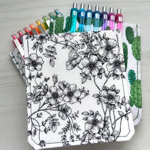 May include: A fabric pen case featuring a white background with a black floral line art pattern, containing a variety of colorful gel pens. Behind it, a second fabric case displays a green cactus print, also filled with multiple pens.