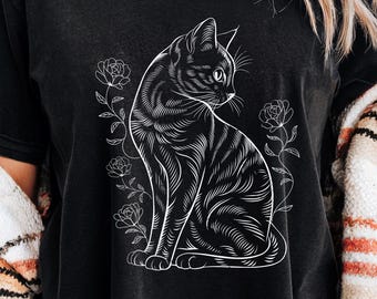 May include: Black t-shirt featuring a white illustration of a cat with floral accents. The cat is depicted in a seated position, with detailed stripes and a focused gaze. The design is centered on the shirt, creating a striking visual.