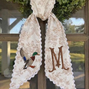May include: A long fabric scarf featuring a cream and tan toile de jouy pattern with scalloped white edges. One end displays an embroidered mallard duck with green, brown, and blue feathers, while the other end features a large, brown embroidered letter M.
