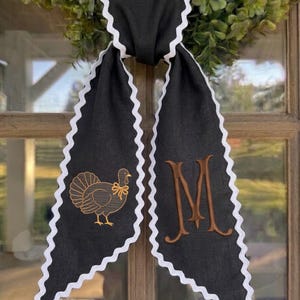 May include: A black fabric ribbon with white rickrack trim along the edges. The left side features an embroidered gold turkey with a small bow, while the right side displays a large, embroidered brown letter M in a serif font style.