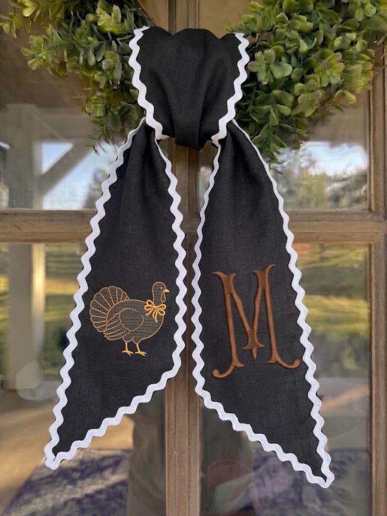 May include: A black fabric ribbon with white rickrack trim along the edges. The left side features an embroidered gold turkey with a small bow, while the right side displays a large, embroidered brown letter M in a serif font style.