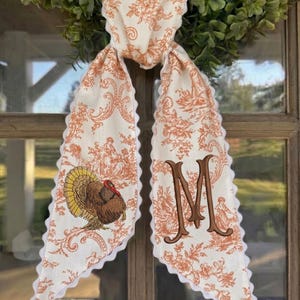 May include: A long fabric scarf featuring an orange and white toile pattern with floral and pastoral motifs. One end displays an embroidered turkey, while the other features a large, brown embroidered letter M. The edges have a white scalloped trim.