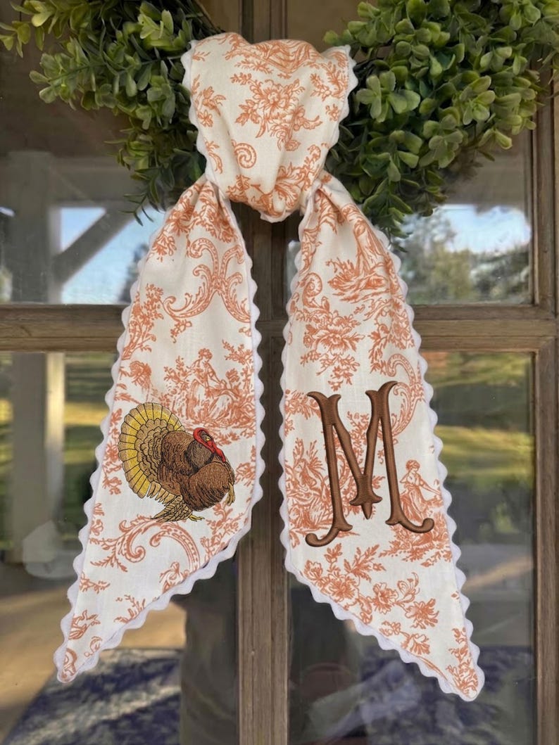 May include: A long fabric scarf featuring an orange and white toile pattern with floral and pastoral motifs. One end displays an embroidered turkey, while the other features a large, brown embroidered letter M. The edges have a white scalloped trim.