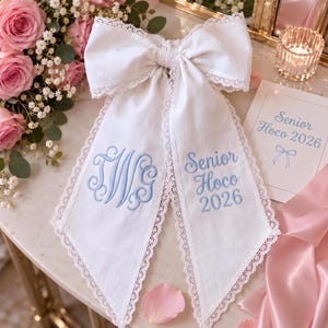 Peut inclure: Un nœud pour cheveux en tissu blanc avec une bordure en dentelle délicate. Le pan gauche présente un monogramme bleu brodé avec les lettres TWG, tandis que le pan droit porte le texte bleu brodé Senior Hoco 2026.